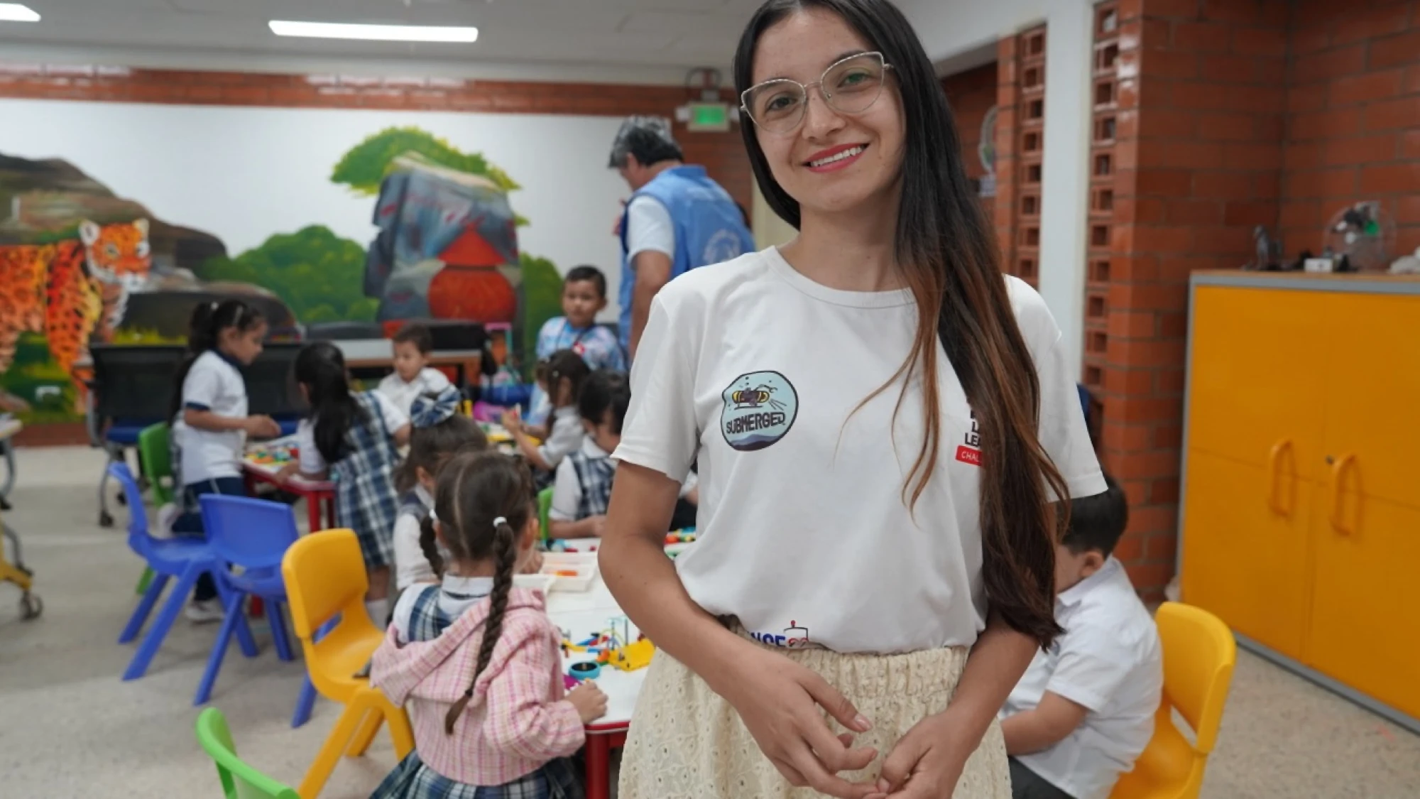 Tati sonriendo en el aula de robótica, con niños armando piezas al fondo